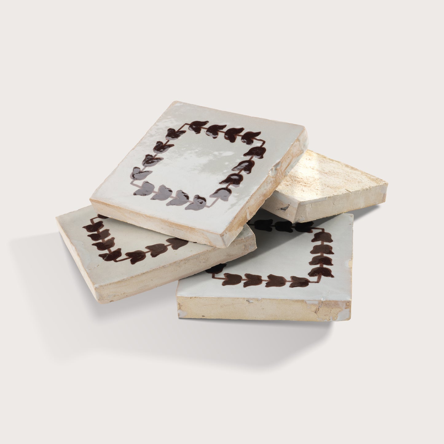 Four square zellige tiles with a distressed off-white glaze, decorated with a dark brown leaf border, arranged in a stack on a light gray background.