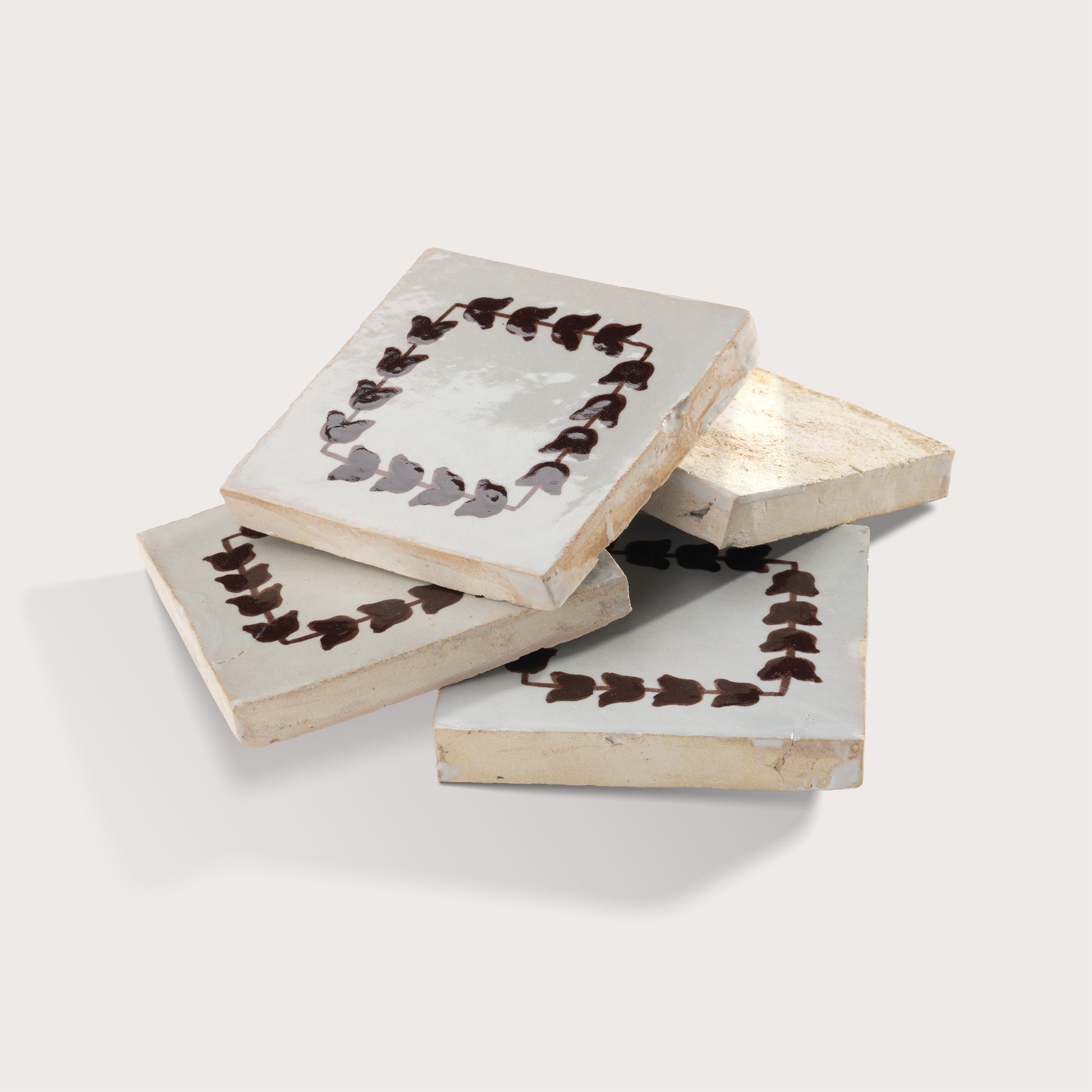 Four square zellige tiles with a distressed off-white glaze, decorated with a dark brown leaf border, arranged in a stack on a light gray background.