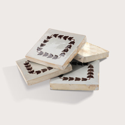 Four square zellige tiles with a distressed off-white glaze, decorated with a dark brown leaf border, arranged in a stack on a light gray background.