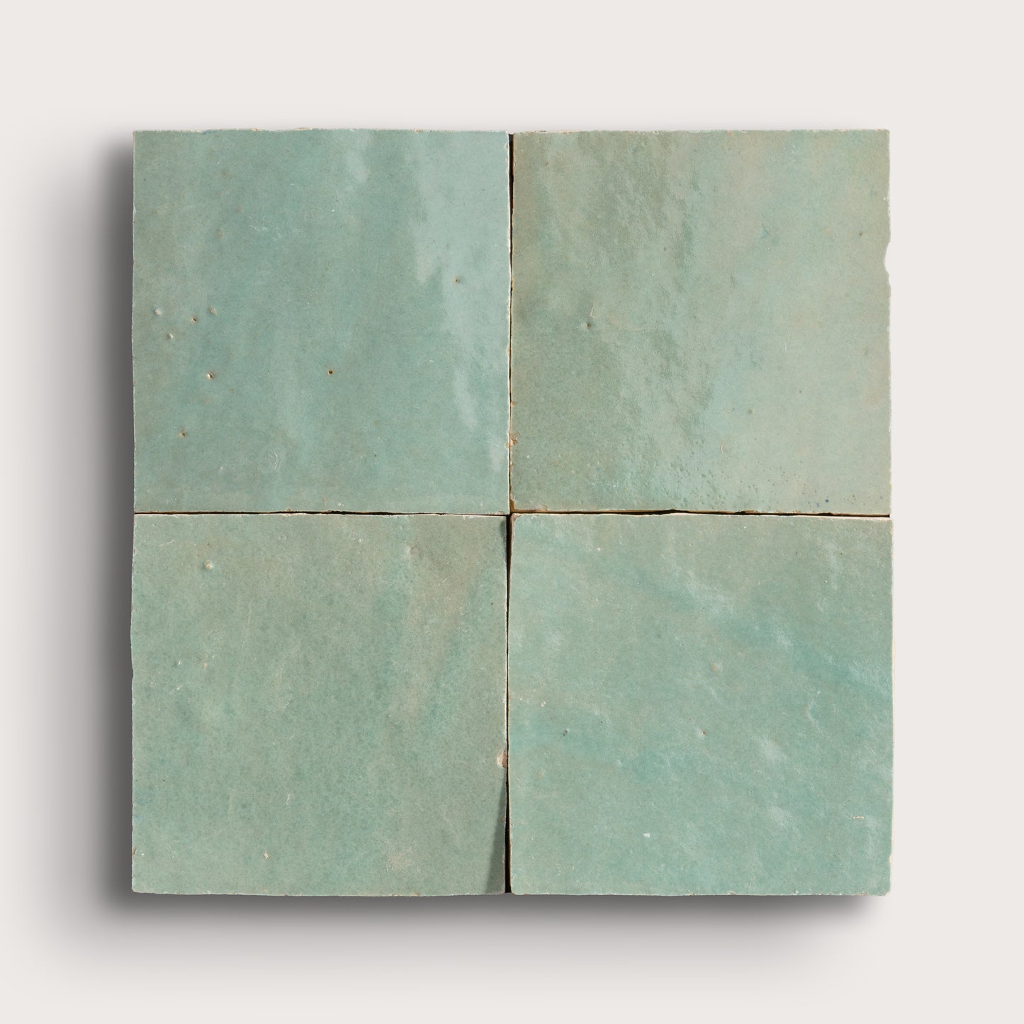 Four square zellige tiles arranged in a 2x2 grid, featuring a textured matte finish in a muted sage green color, set against a light gray background.