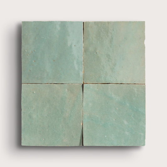 Four square zellige tiles arranged in a 2x2 grid, featuring a textured matte finish in a muted sage green color, set against a light gray background.