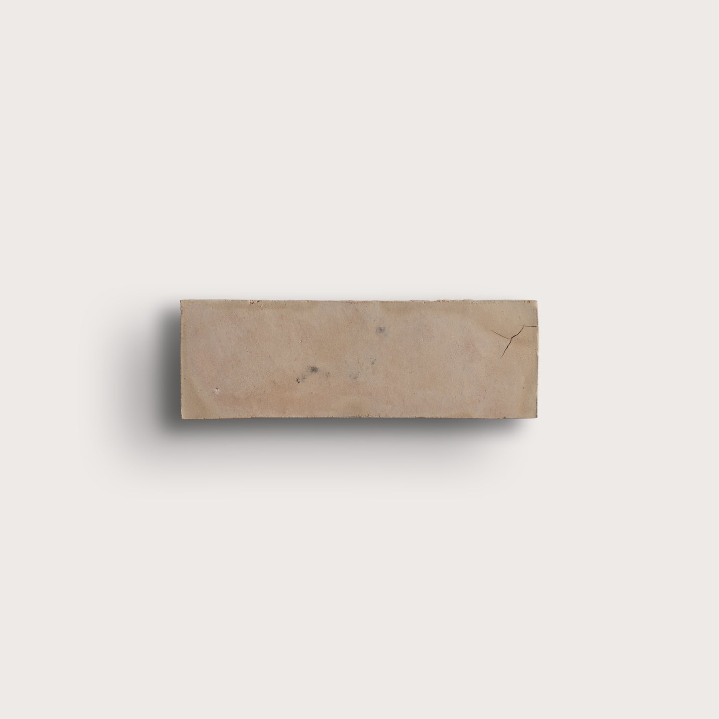 A rectangular beige zellige tile with natural marbling and a small crack, shown against a solid off-white background with a soft drop shadow.