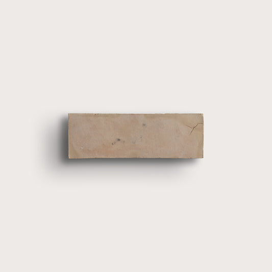 A rectangular beige zellige tile with natural marbling and a small crack, shown against a solid off-white background with a soft drop shadow.