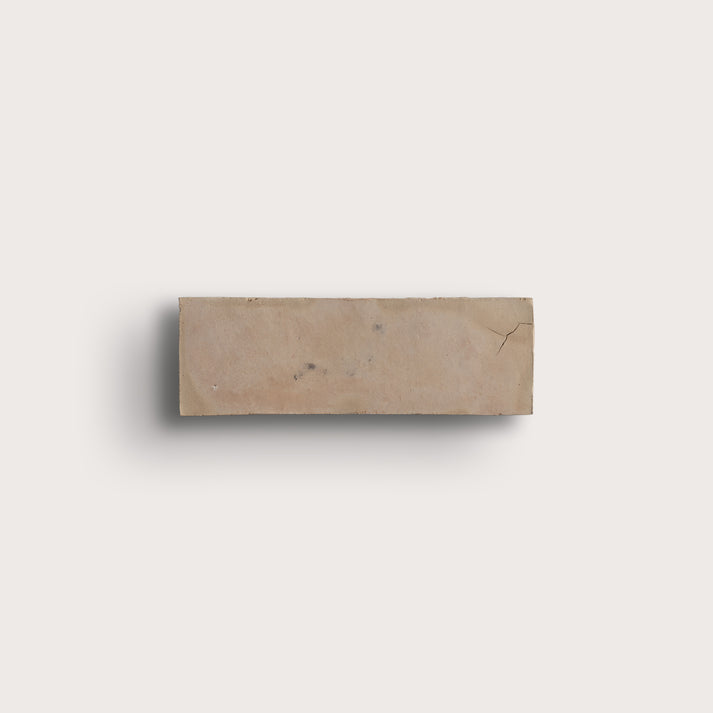 A rectangular beige zellige tile with natural marbling and a small crack, shown against a solid off-white background with a soft drop shadow.