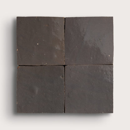 Four square dark charcoal gray zellige tiles arranged in a 2x2 grid, showing a matte textured surface with subtle natural variations, set against a light gray background.