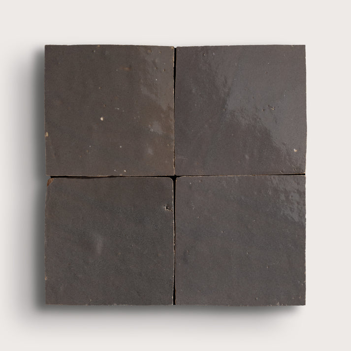 Four square dark charcoal gray zellige tiles arranged in a 2x2 grid, showing a matte textured surface with subtle natural variations, set against a light gray background.