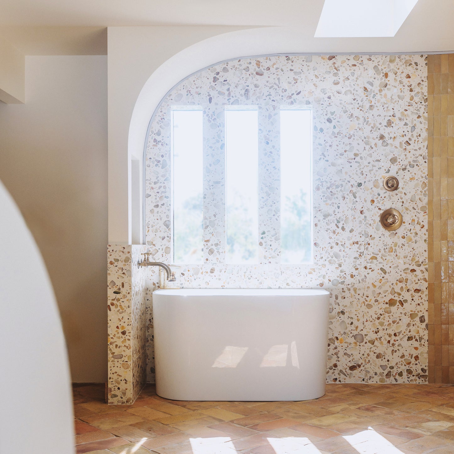 A modern freestanding white bathtub placed in a bathroom with terrazzo tile walls and herringbone tile flooring, lit by natural light from an arched window.