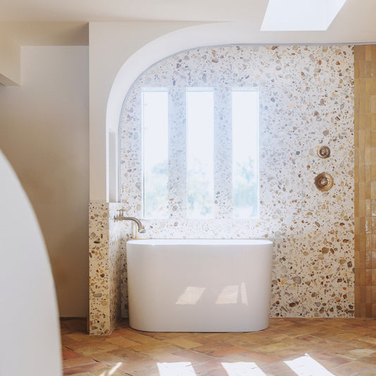 A modern freestanding white bathtub placed in a bathroom with terrazzo tile walls and herringbone tile flooring, lit by natural light from an arched window.