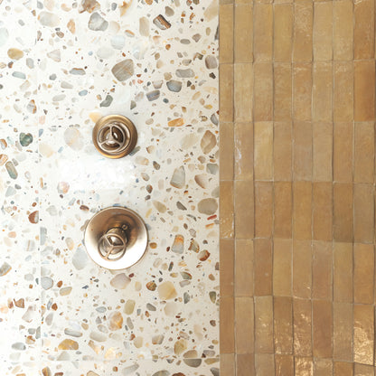 Close-up of a bathroom tile and fixture area showing a white terrazzo wall with multicolored flecks, a vertical band of warm tan rectangular tiles, and two round metallic shower controls.