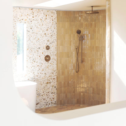 A modern shower stall with warm beige stacked tile walls and floor, a white textured terrazzo-patterned wall panel, and a brass shower head and controls, shown in a bright bathroom interior.