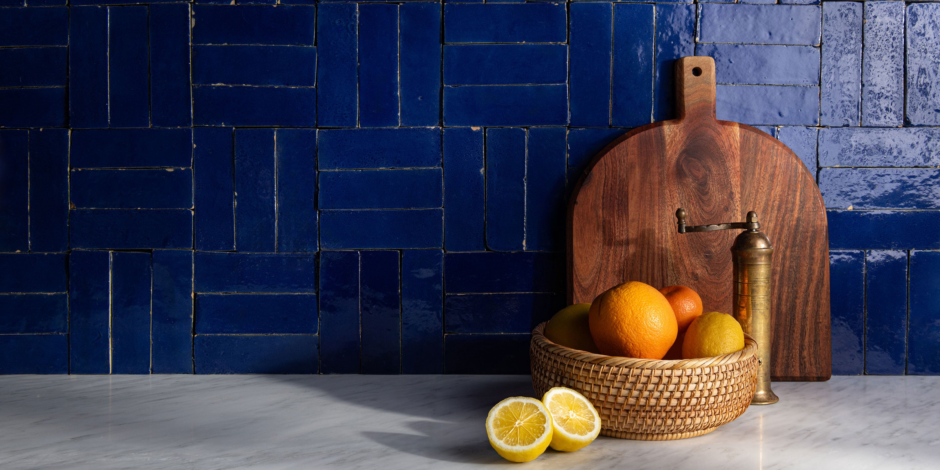 Wooden cutting board and basket with fruits against a blue zellige tiled wall