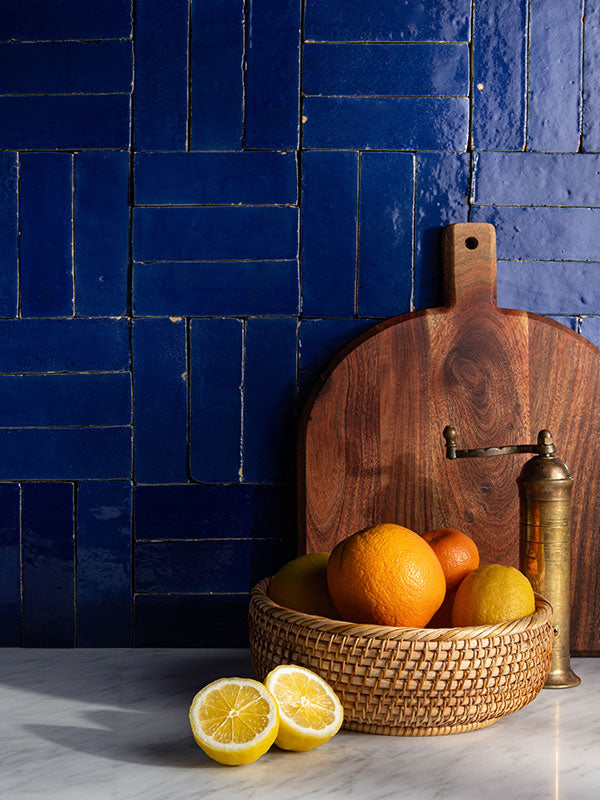 Wooden cutting board with a basket of fruits against a blue zellige tiled wall.