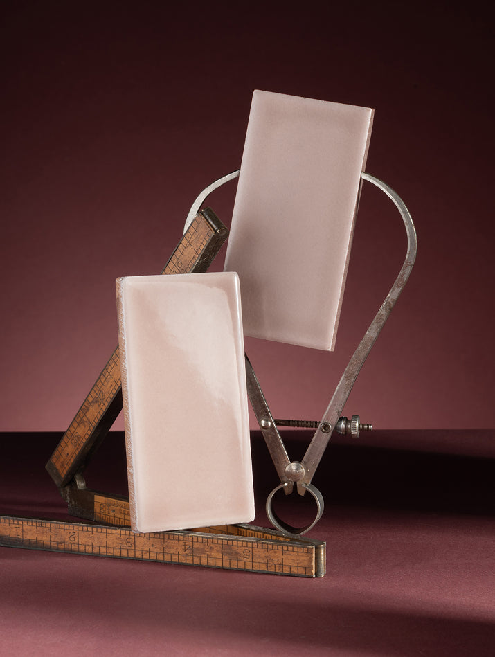 Two rectangular pale pink ceramic tiles displayed on a vintage metal and wood measuring device against a dark burgundy background.