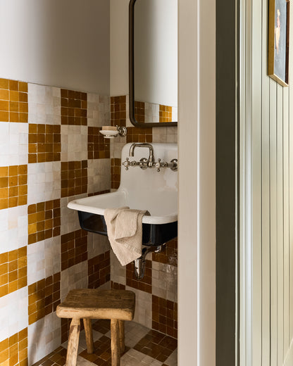 Bathroom with white sink, wooden stool, and yellow and brown zellige tiled walls.
