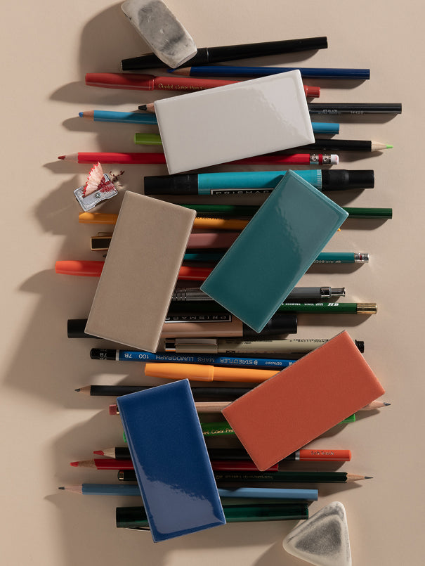A styled flat-lay of various colored pencil erasers in different colors (blue, teal, orange, beige, white) arranged on top of a pile of colored pencils on a warm beige background.
