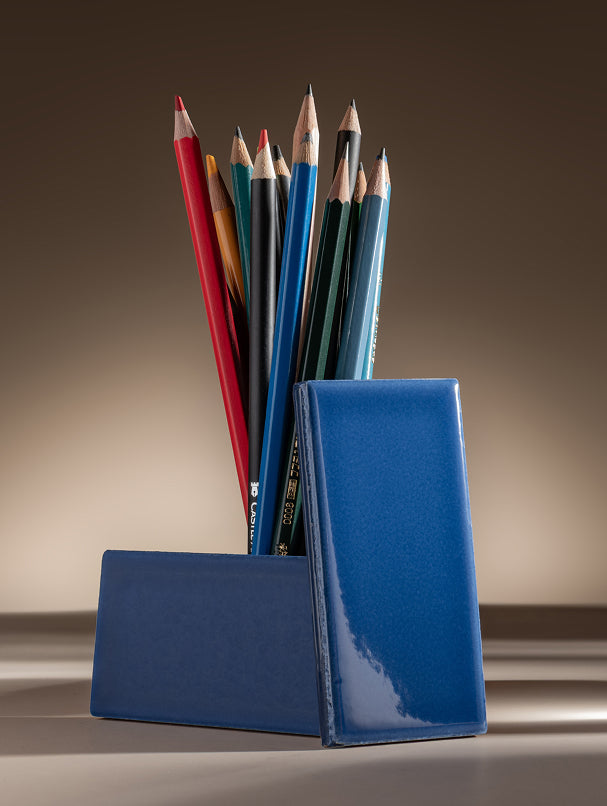 A glossy ceramic tile, holding a fan arrangement of colored pencils, photographed on a warm beige studio background.