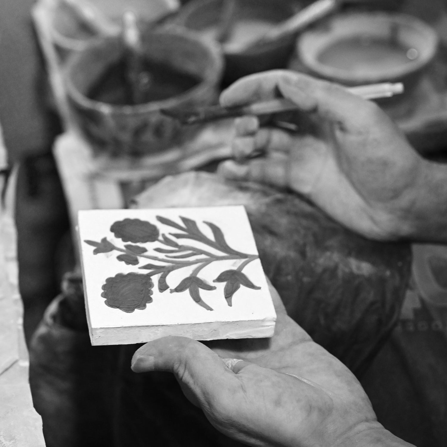 A black and white close-up of a person's hands holding a small square tile or block printed with a floral design, with a blurred workshop background containing bowls and tools.