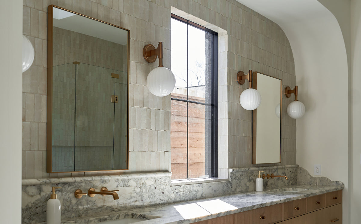 A double vanity bathroom scene with two large rectangular mirrors, round white globe wall sconces, brass faucets, a marble countertop, and a window looking out onto a wooden fence.