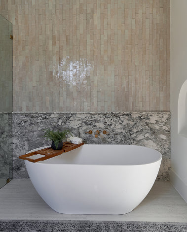 A modern freestanding oval bathtub in white, set against a beige mosaic tile wall and a gray marble band, with a wooden tray draped over the tub edge holding a small plant and folded towels.
